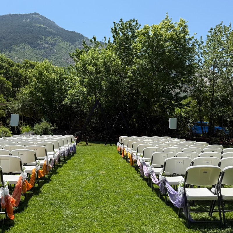Folding chairs with colorful sashes on grass facing trees and mountains in the background.