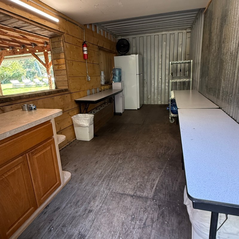 Small room with wooden and metal walls, a fridge, tables, and a fire extinguisher.
