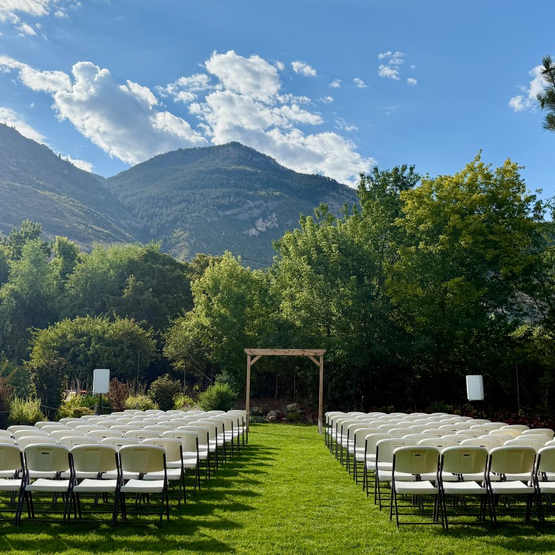 Outdoor wedding setup with chairs and arch, mountains in background, sunny sky.