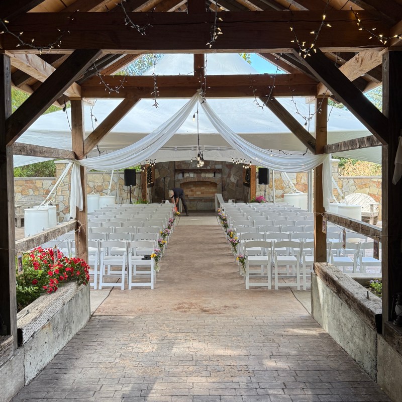Outdoor wedding venue with white chairs, draped fabric, string lights, and floral decorations.