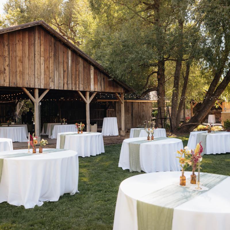 Outdoor wedding setup with round tables, white tablecloths, and floral centerpieces near a wooden pavilion.
