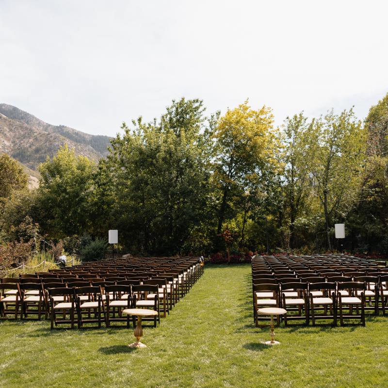 Outdoor wedding setup with rows of empty chairs on a grassy area, surrounded by trees and mountains.
