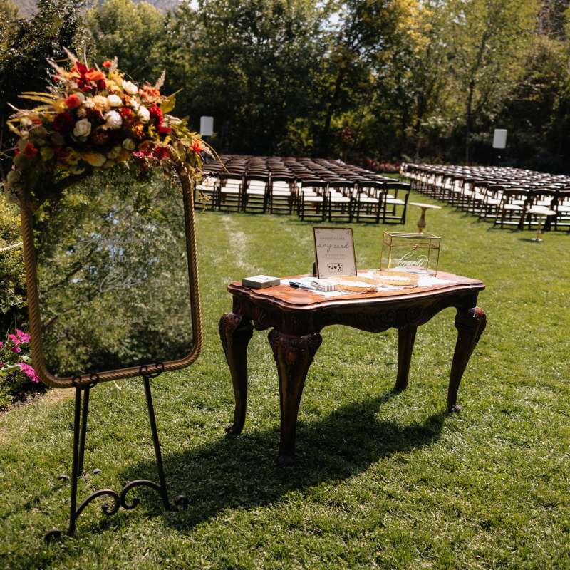 Outdoor wedding setup with decorated mirror, antique table, and rows of chairs on a grassy lawn.