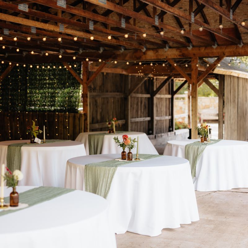 Wedding venue with round tables, white cloths, green runners, flower vases, and string lights in a rustic setting.